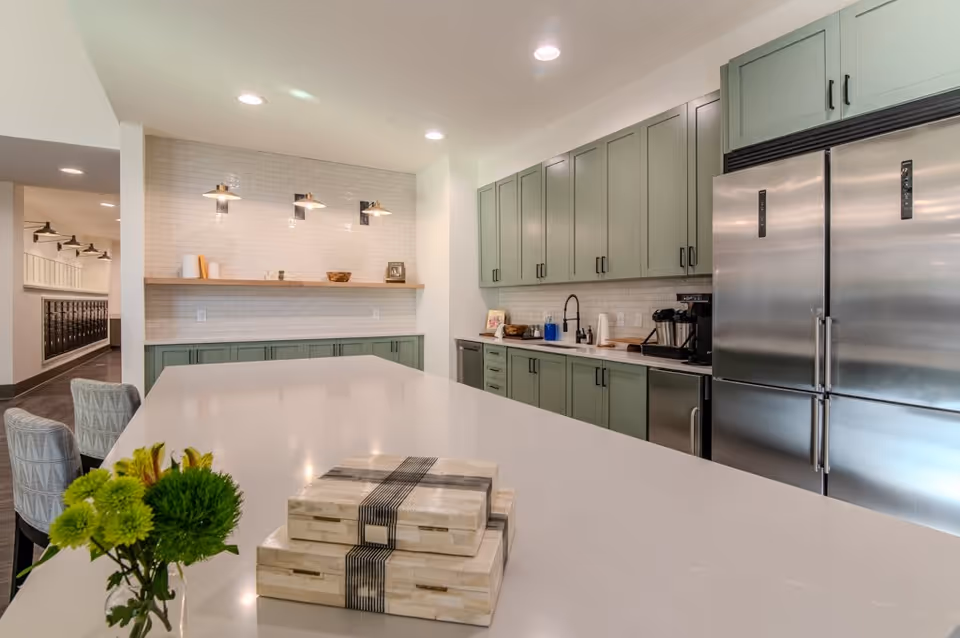 Modern kitchen area with a large white island countertop, green cabinetry, stainless steel double-door refrigerator, and a coffee machine. There are three wall-mounted lights above a wooden shelf on a white tiled backsplash. Two patterned chairs are partially visible on the left side of the island, and a small green floral arrangement and decorative boxes are on the countertop.