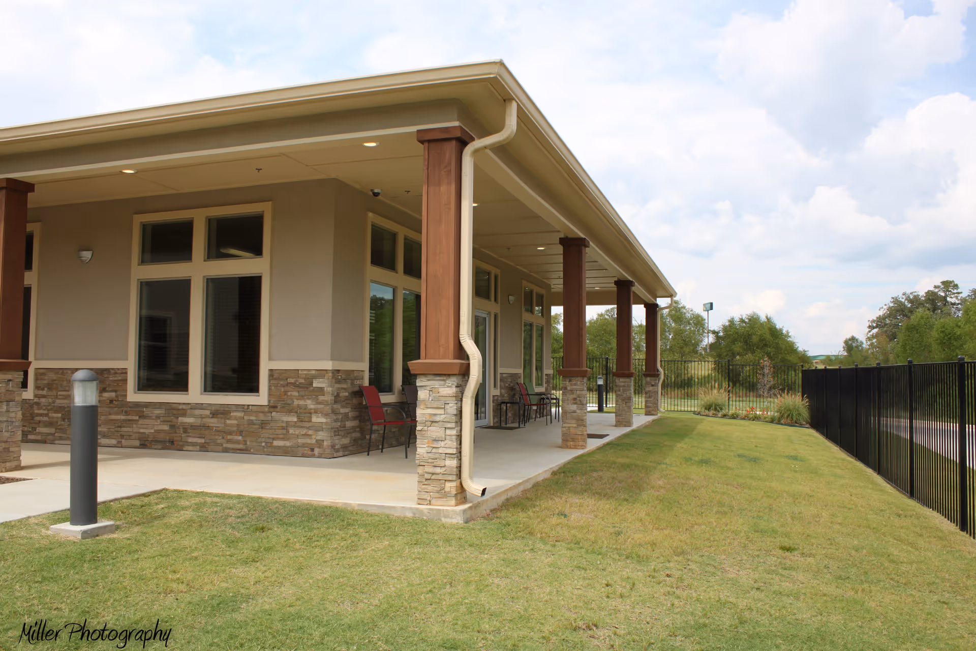 Exterior view of a building with a covered patio supported by stone and wood columns. The patio has several chairs and overlooks a grassy area with a black metal fence. The sky is partly cloudy.