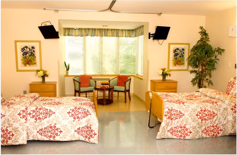 A bright and clean room with two beds covered in red and white patterned bedspreads. Between the beds are two wooden nightstands with flower arrangements. At the far end of the room is a large window with blinds and a valance, flanked by two chairs with red cushions and a small round table with books. There are framed botanical prints on the walls and a tall green plant in the corner.
