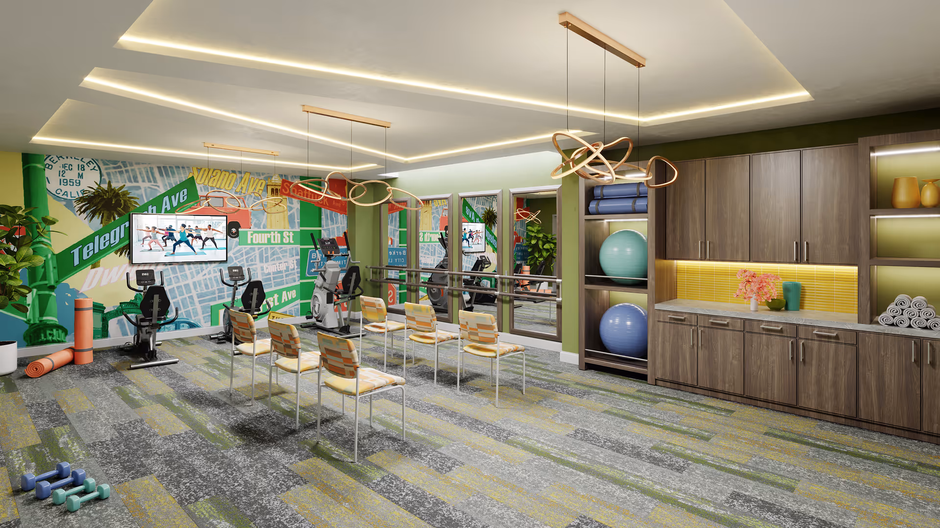 A bright and modern exercise room with colorful wall art depicting street signs and a map. The room features exercise bikes, chairs arranged in rows, large mirrors on the wall, shelves with exercise balls and rolled towels, and contemporary pendant lighting. The floor has a patterned carpet in shades of gray and green.