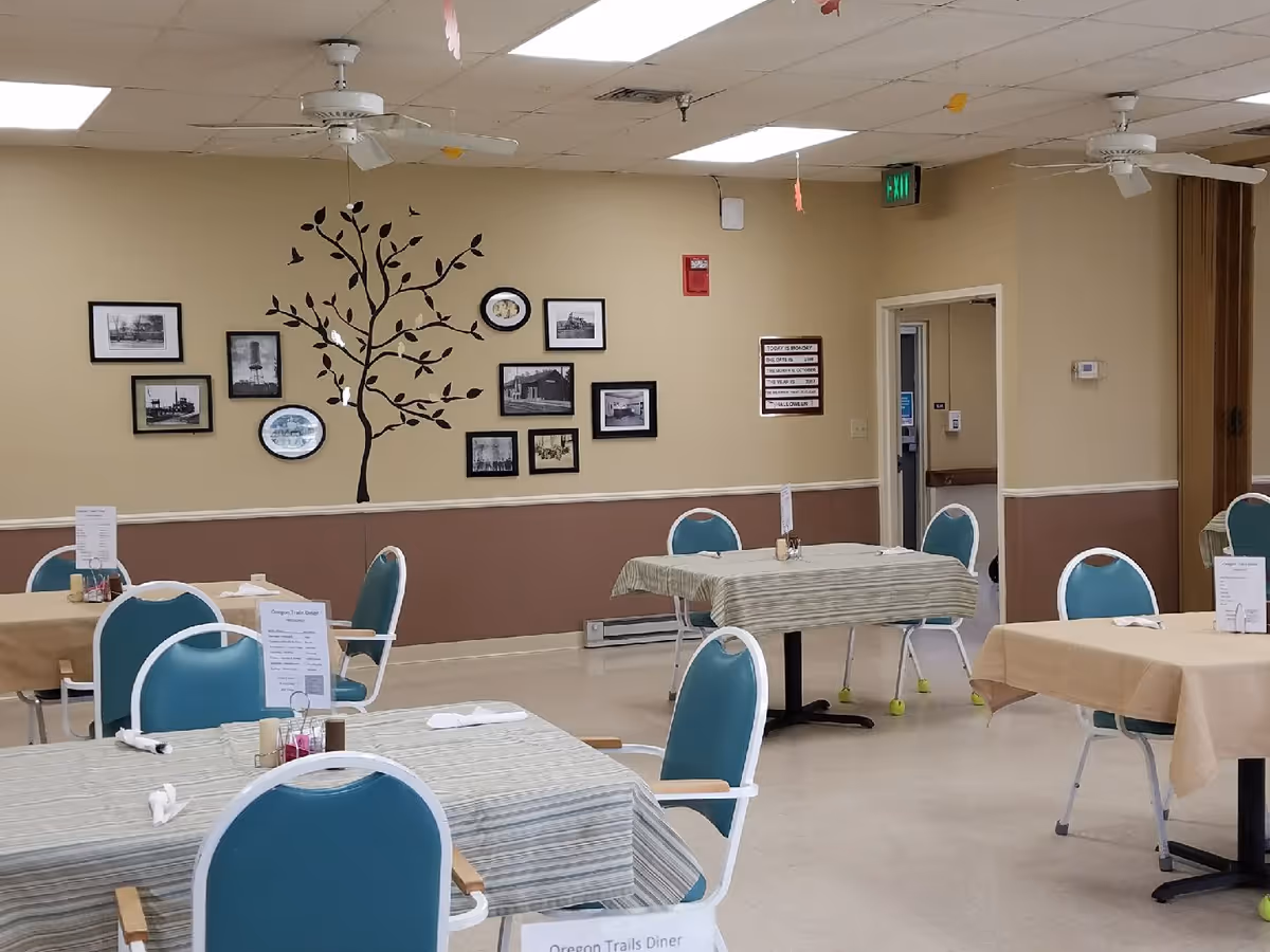 A dining room with several tables covered with tablecloths and set with menus, napkins, and condiments. Blue chairs with white frames surround the tables. The walls are beige with a brown lower half and decorated with framed black and white photos and a tree wall decal. Ceiling fans and an exit sign are visible on the ceiling.
