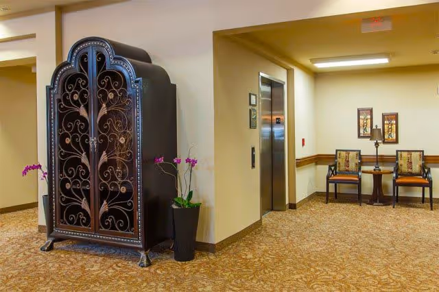 Interior hallway area of a senior living facility with a decorative black metal cabinet on the left, a potted plant with purple flowers beside it, an elevator with closed doors in the center, and two chairs with a small table and lamp between them on the right. The walls are beige and the floor is carpeted with a patterned design.