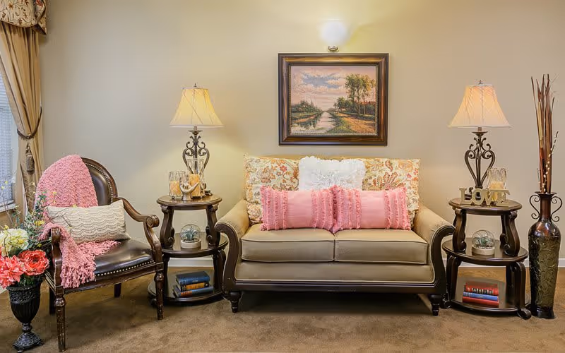 A cozy living room area with a beige sofa adorned with floral and pink pillows, flanked by two wooden side tables each holding a decorative lamp. To the left, there is a wooden armchair with a pink throw blanket and a white pillow. A framed landscape painting hangs on the wall above the sofa. A vase with flowers is placed on the floor to the left, and a tall vase with decorative sticks is on the right side table.
