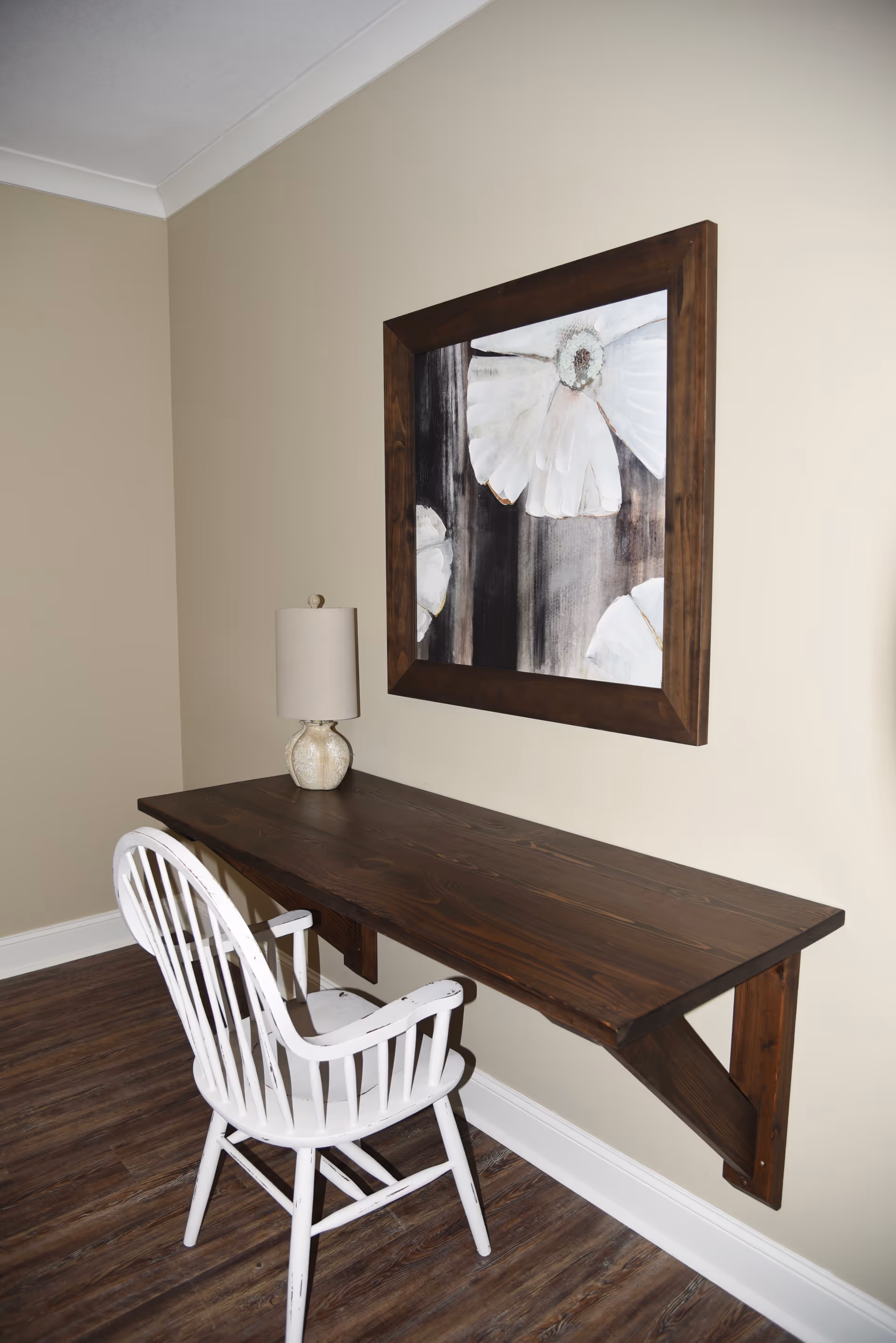 A small workspace with a dark wooden wall-mounted desk and a white wooden chair. On the desk is a beige table lamp with a textured base. Above the desk hangs a large framed painting of white flowers on a dark background. The room has beige walls and dark wood flooring.