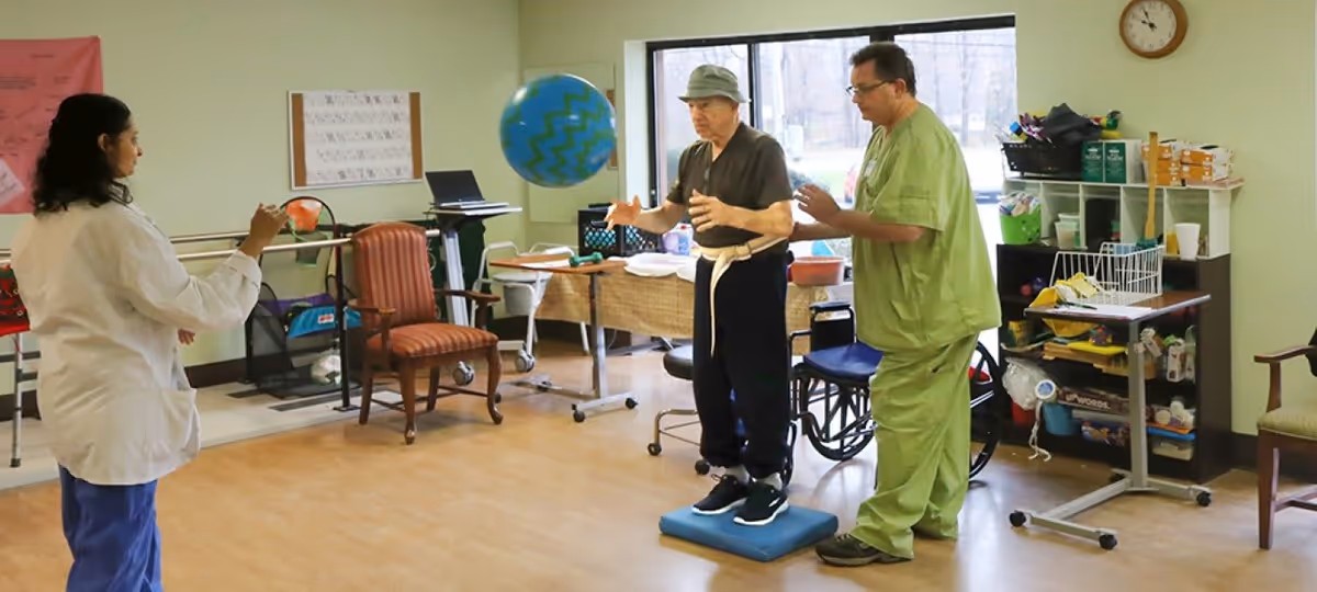 An elderly man wearing a hat stands on a blue balance pad while catching a large blue ball with green zigzag patterns. He is assisted by a man in green scrubs, and a woman in a white coat stands opposite them, appearing to throw the ball. The room has wooden flooring, chairs, a wheelchair, and various medical and therapy equipment, with a large window in the background.