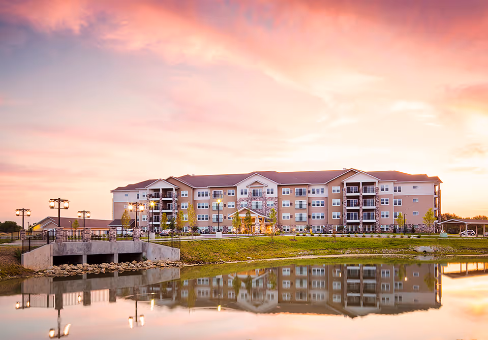 A large, multi-story senior living apartment building named Oxford Villa Active Senior Apartments is shown at sunset with a colorful sky. The building is reflected in a calm pond in the foreground, and there are street lamps and a small bridge near the water.