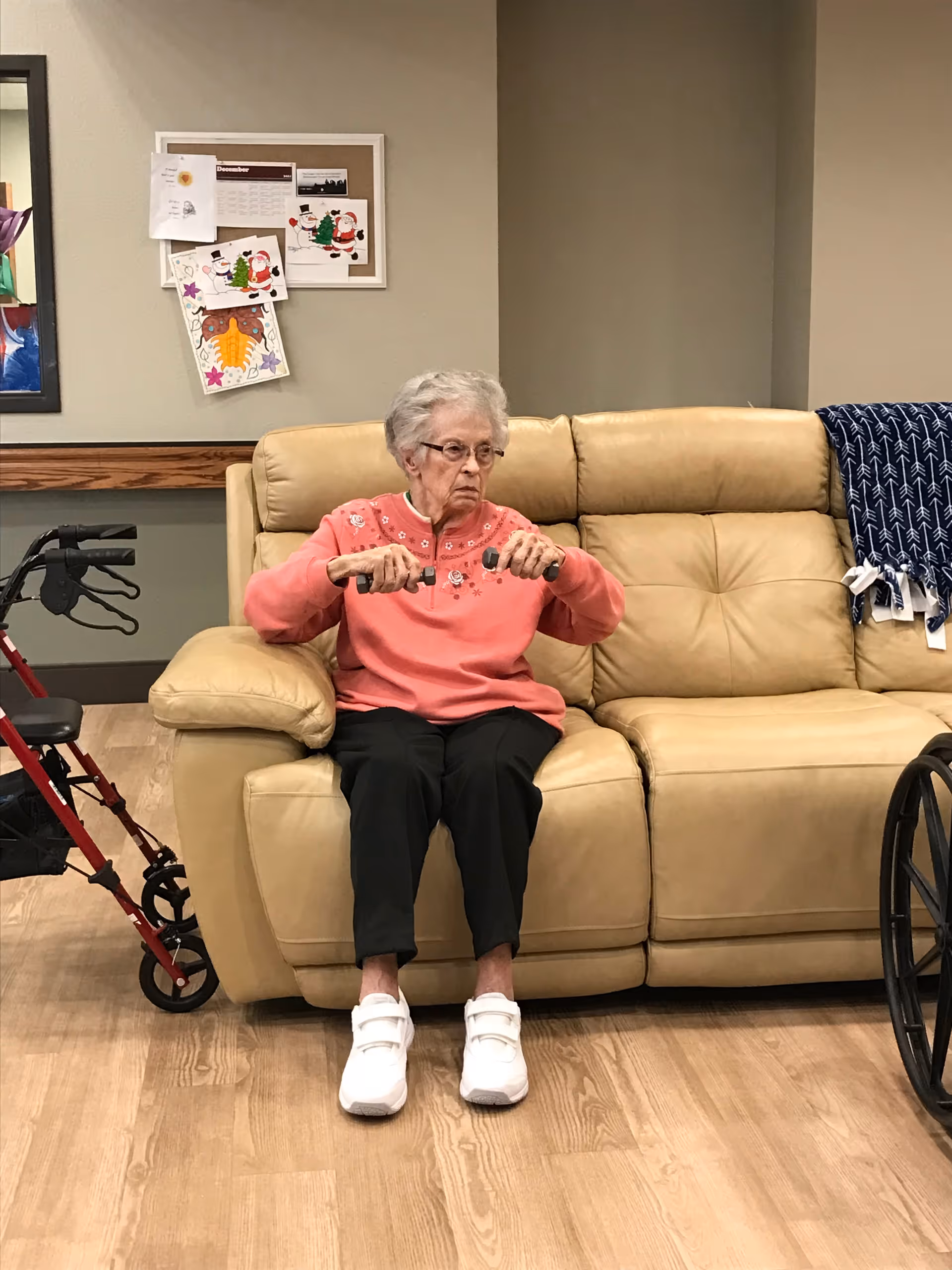 An elderly woman wearing a pink sweater and black pants is sitting on a beige leather couch in a room. She is holding small hand weights and appears to be exercising. To her left is a red walker, and to her right is a wheelchair. Behind her on the wall is a bulletin board with holiday-themed decorations and papers.