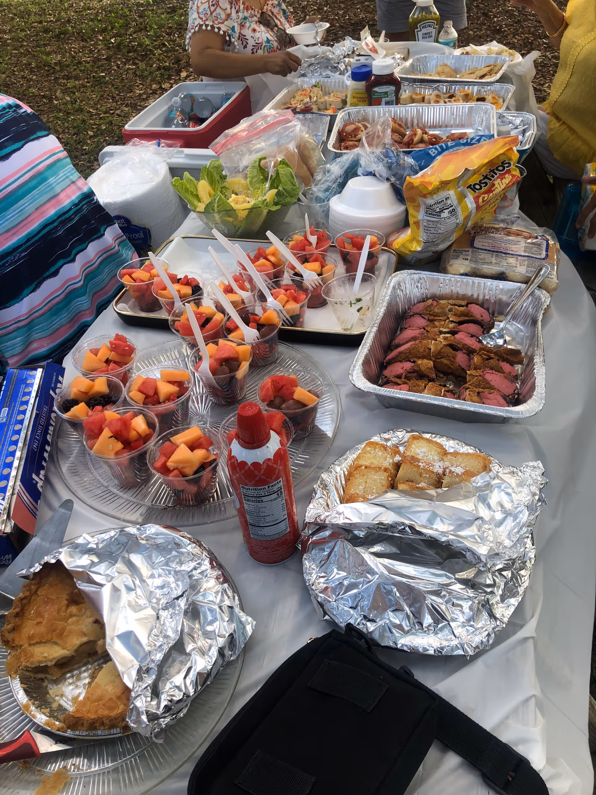 Outdoor buffet table with trays of fruit cups, sliced meat, foil-covered dishes, chips and condiments, with people serving themselves.