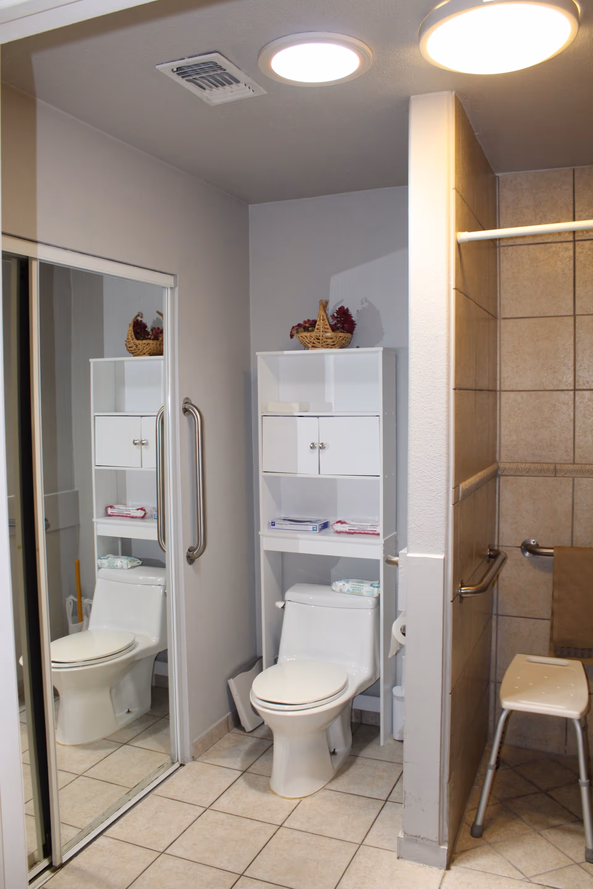 Bathroom with a toilet beneath a white over-toilet shelving unit, mirrored closet door, grab bars, tiled shower with a shower stool, and ceiling lights.