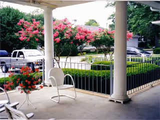 Covered front porch with white columns and metal chairs overlooking pink-flowering trees, trimmed hedges, and parked cars.