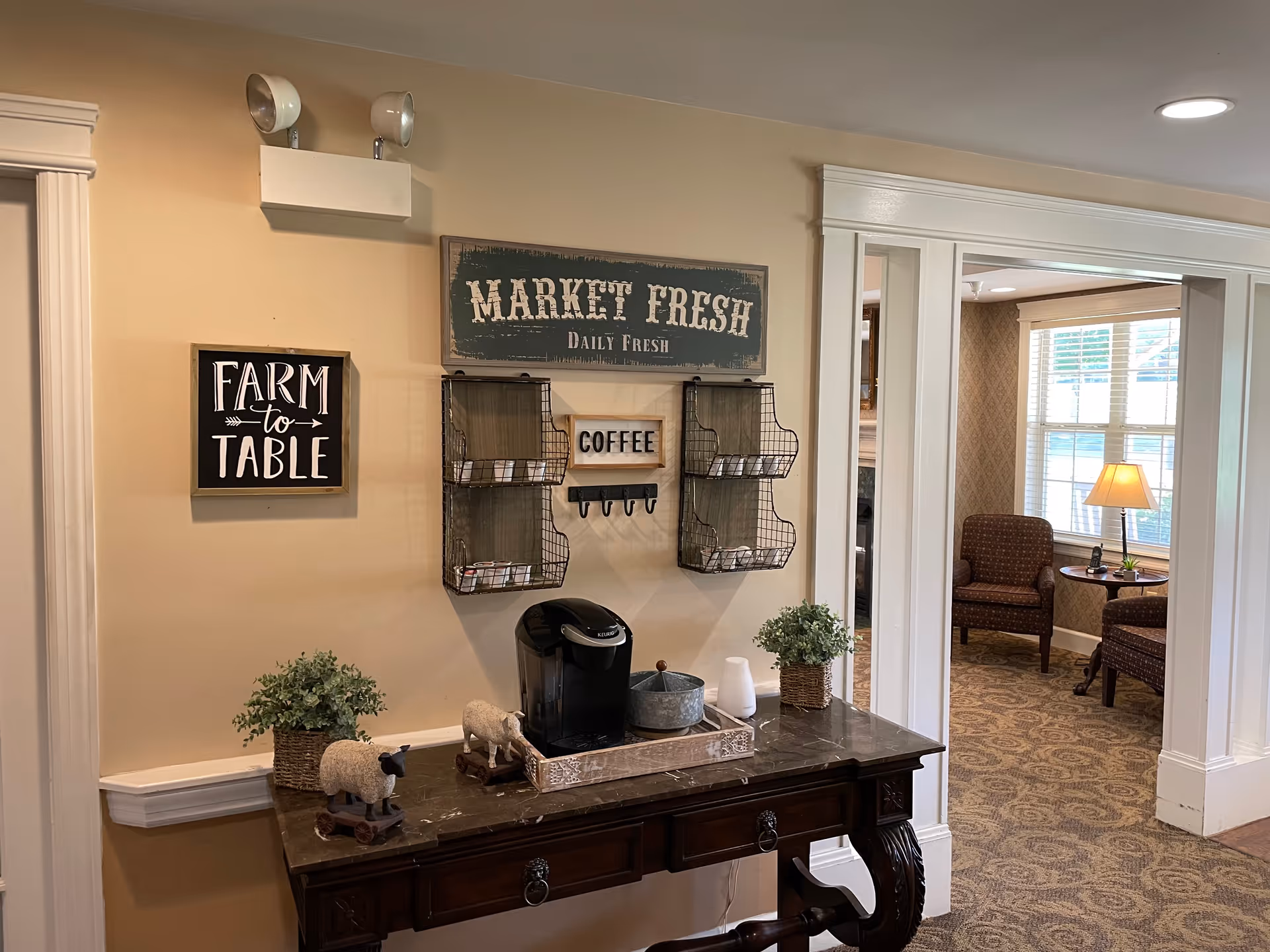 A cozy interior corner with a dark wooden table holding a coffee maker, decorative sheep figurines, and potted plants. Above the table are wall decorations including a sign that reads 'MARKET FRESH Daily Fresh', a smaller sign saying 'COFFEE', and another sign that says 'FARM to TABLE'. In the background, there is a sitting area with two upholstered chairs, a small round table with a lamp, and a window with blinds.