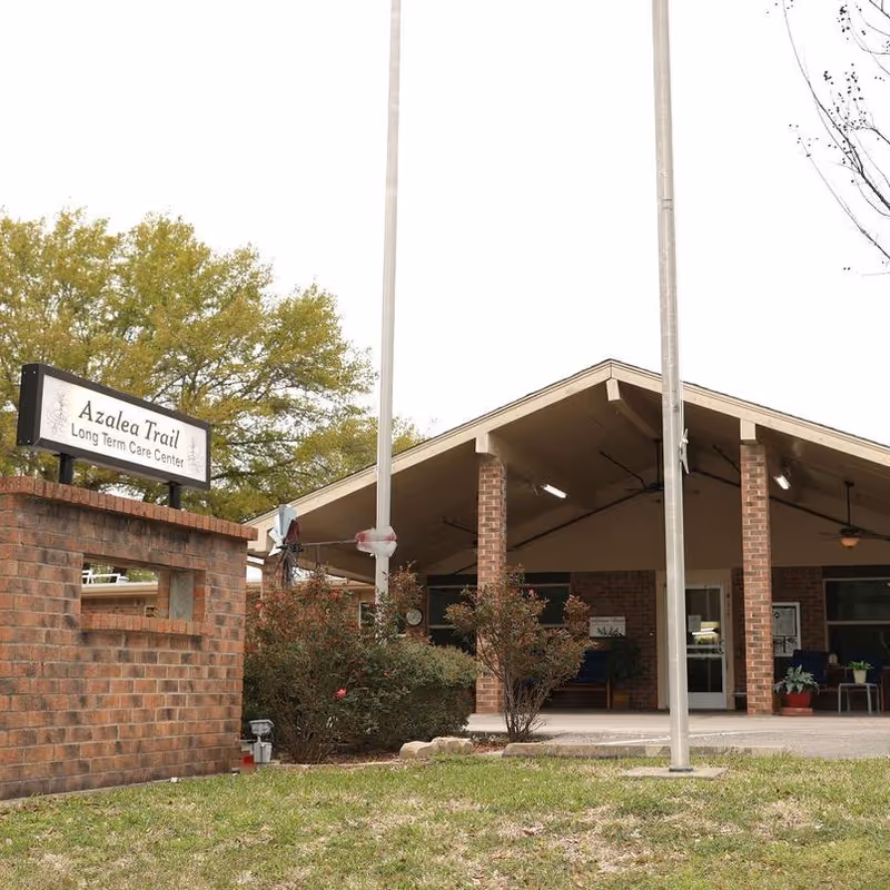 Exterior view of Azalea Trail Long Term Care Center showing a brick entrance sign, two flagpoles, and a covered porch area with brick pillars and some outdoor seating. Trees and shrubs are visible around the building.