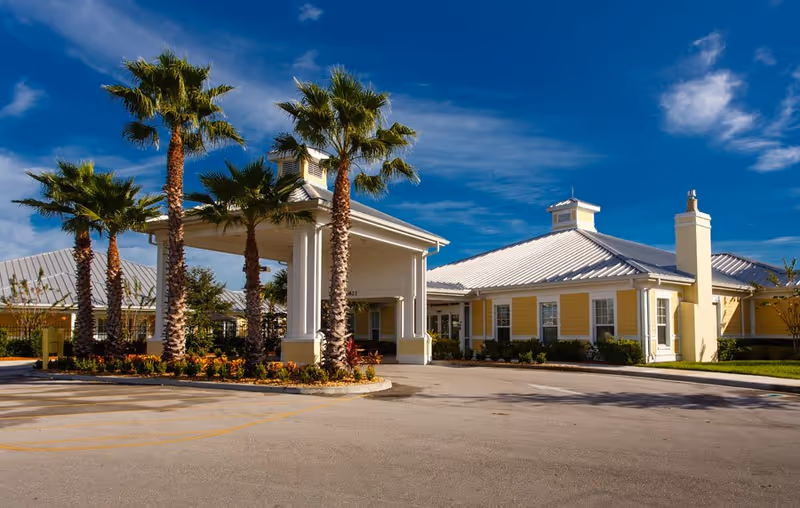 Exterior view of The Brennity at Tradition Senior Living facility showing a yellow building with a metal roof, a covered entrance supported by columns, palm trees, and a clear blue sky.