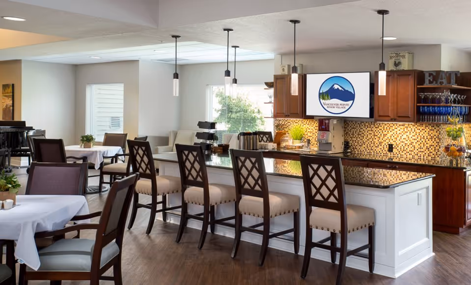 Communal dining area with a kitchen-style bar, high stools, dining tables, and a TV showing the Vancouver Pointe logo.