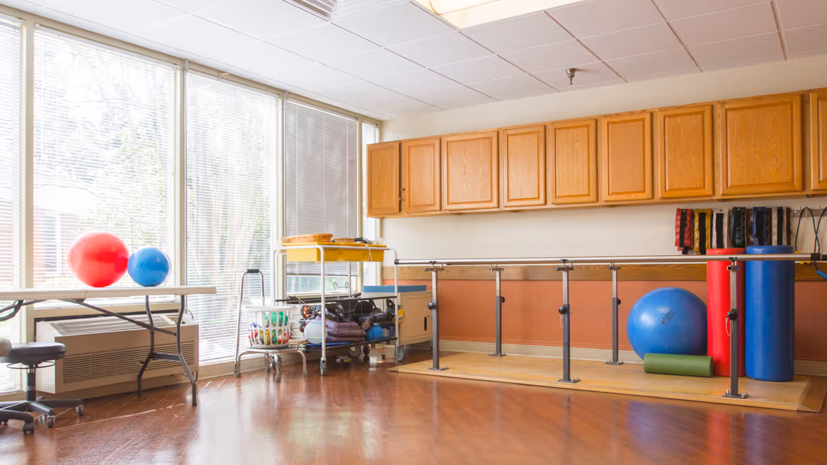 A rehabilitation therapy room with large windows covered by blinds, wooden cabinets mounted on the wall, parallel bars for walking exercises, and various exercise equipment including large red and blue therapy balls, foam rollers, and resistance bands. There is also a table with a chair and a cart holding additional therapy supplies.