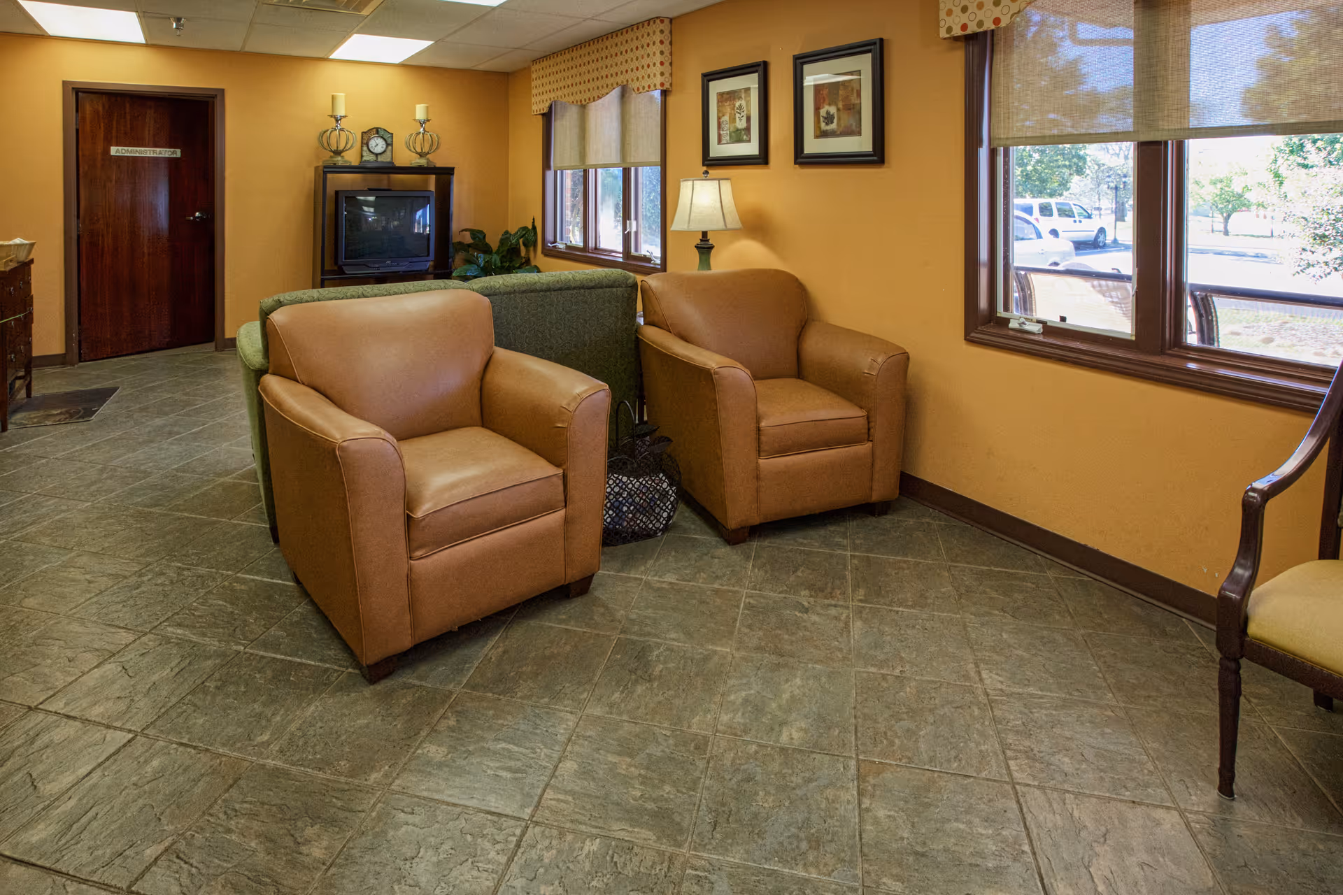 A cozy interior room with two brown leather armchairs and a green couch arranged around a TV on a stand. The walls are painted a warm yellow, decorated with framed pictures and a table lamp. There are two large windows with beige blinds letting in natural light, and a door labeled 'ADMINISTRATIVE' is visible in the background.