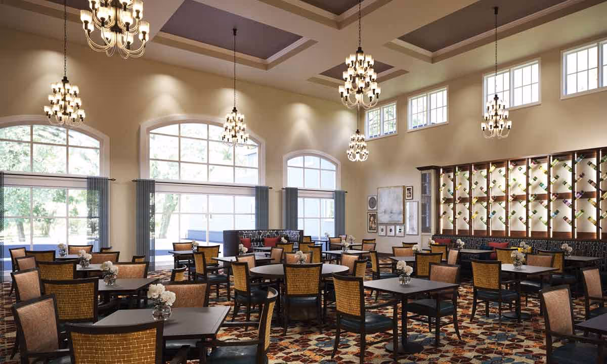A spacious dining room with multiple square tables and chairs arranged neatly. The room features large arched windows with blue curtains, several elegant chandeliers hanging from a high ceiling with recessed panels, and a decorative wall display with bottles. The floor has a colorful patterned carpet.