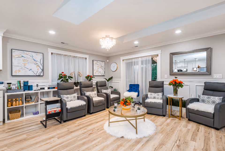 Bright communal living room with gray recliners arranged around a round glass coffee table, plants and decorative accents.