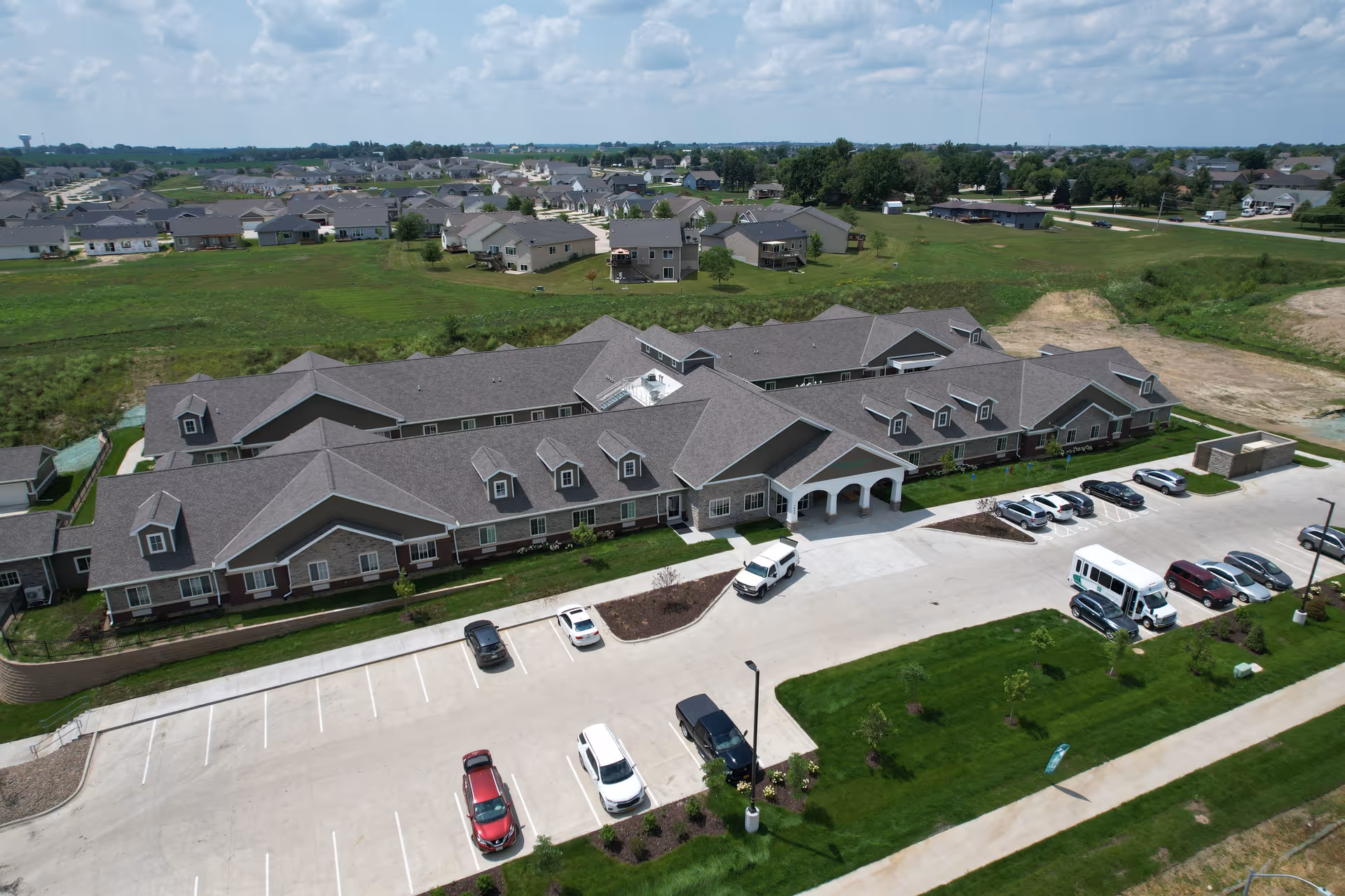 Aerial view of Edencrest at The Tuscany, a large senior living facility with a gray roof and stone facade. The building is surrounded by green lawns and a parking lot with several cars and a shuttle bus. In the background, there are residential houses and open fields under a partly cloudy sky.