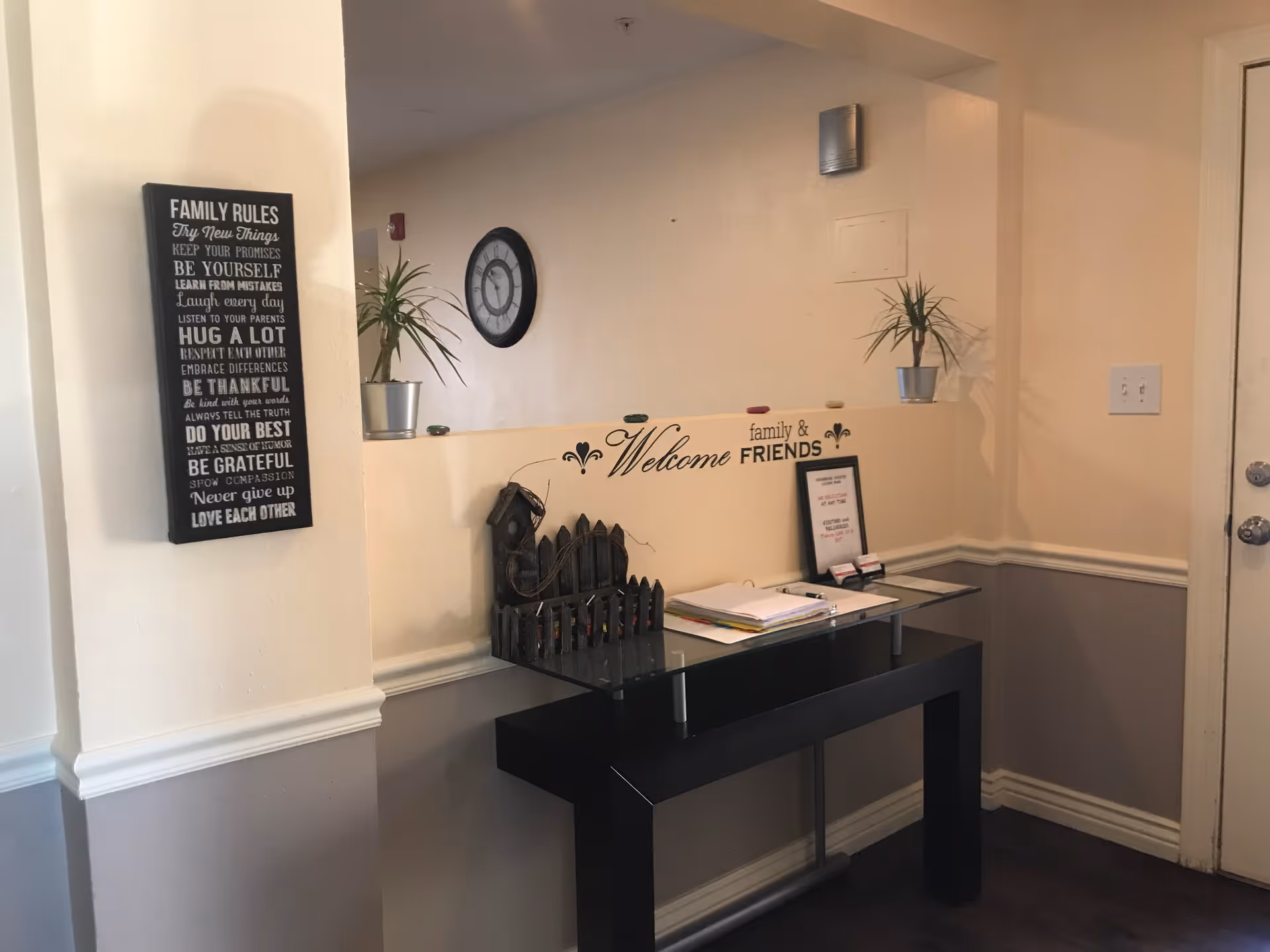 Interior corner of a room with a black glass-top table against the wall. On the wall above the table, there is a decorative sign that says 'Welcome family & FRIENDS'. Two small potted plants are placed on the ledge above the table. A black wall clock is mounted on the wall in the background. To the left, a framed sign with family rules is hanging on the wall. The room has beige and gray walls with white trim.