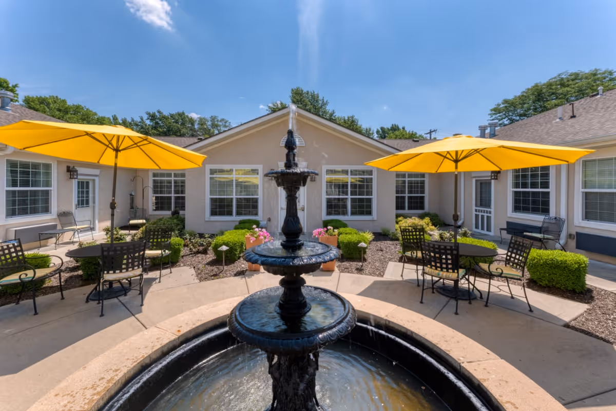 Outdoor courtyard area with a black three-tiered water fountain in the center, surrounded by concrete pathways, green bushes, and flower pots. There are two yellow patio umbrellas shading metal tables and chairs. The courtyard is enclosed by a beige building with multiple windows and doors under a clear blue sky.