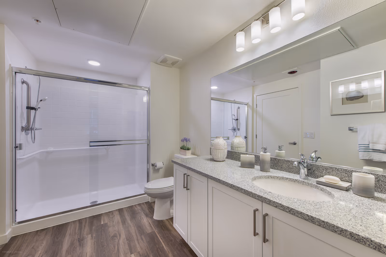 A modern bathroom with a large glass-enclosed shower, a toilet, and a long granite countertop with a sink. The countertop has decorative items including two vases, soap dispensers, and a bar of soap. A large mirror with three light fixtures is mounted above the sink. The floor has wood-style flooring and the walls are painted white.