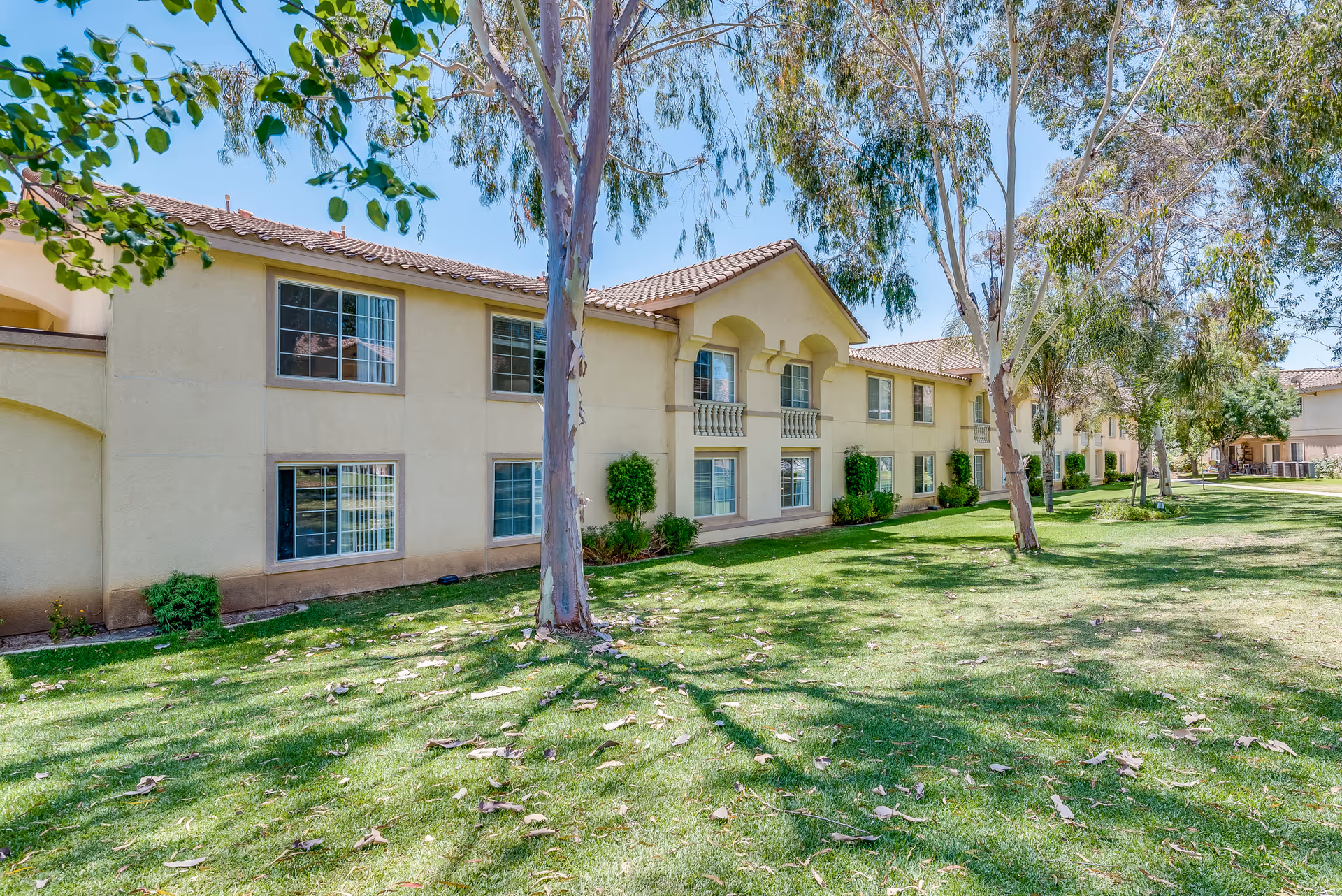 Two-story beige senior living building with a tiled roof facing a grassy lawn and trees.