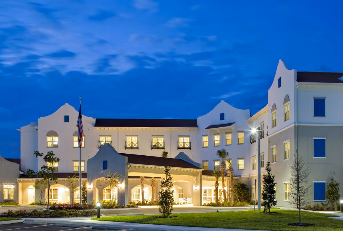 Exterior view of a large, well-lit senior living facility building at dusk with an American flag in front and landscaped greenery around the entrance.