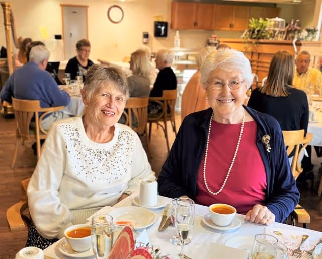 Two elderly women sitting at a dining table in a communal dining room, smiling at the camera. The table is set with cups of tea, glasses of water, and plates. Other seniors are seated at tables in the background, engaged in conversation.