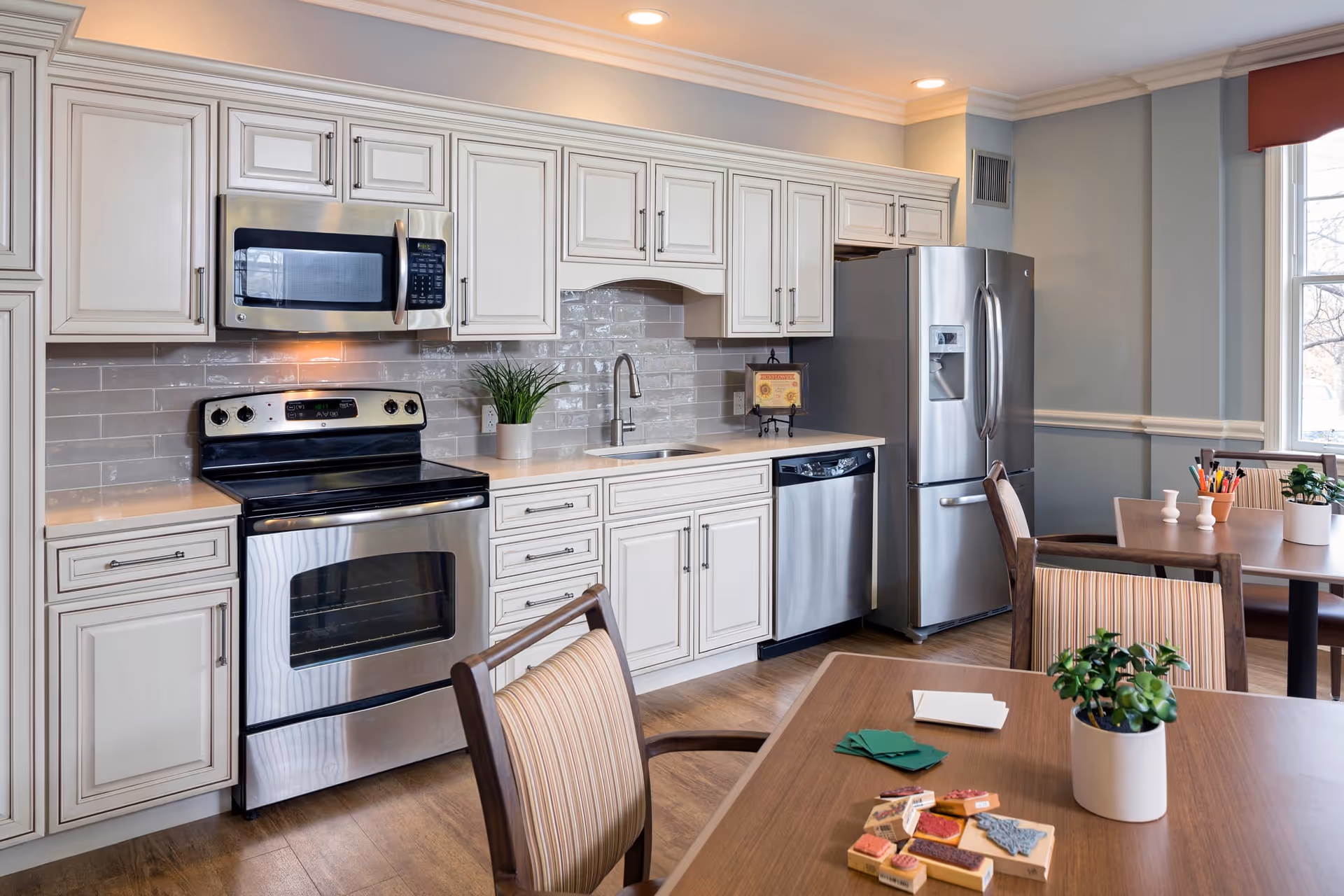 A modern kitchen area with white cabinetry, stainless steel appliances including a microwave, oven, dishwasher, and refrigerator. There are two wooden tables with striped cushioned chairs, small potted plants, and some cards and game pieces on the table. The room has light gray walls and a window letting in natural light.