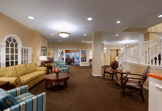 A spacious senior living facility common area with beige walls and brown carpet. The room features a yellow sofa, two striped armchairs, a round wooden coffee table with a flower arrangement, and two wooden chairs with leather seats around a small round table. There is a white staircase with a railing on the right side and a dining area visible through glass doors in the background. The lighting is warm with ceiling lights and a hanging light fixture.