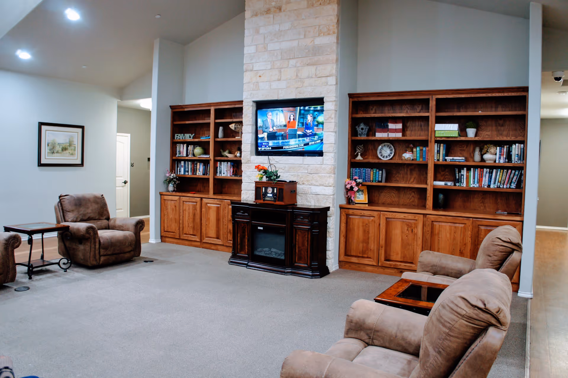 A cozy senior living common area with beige carpet, two brown recliner chairs, a small wooden side table, and built-in wooden bookshelves filled with books and decorative items. A flat-screen TV is mounted on a stone fireplace in the center of the room.