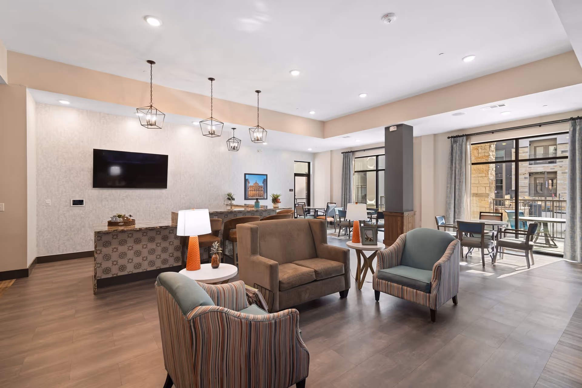 A modern and spacious living room area in ArborView Circle C Ranch featuring a brown loveseat, two striped armchairs, and two small round tables with orange lamps. Behind the seating area is a counter with bar stools and three pendant lights hanging from the ceiling. A flat-screen TV is mounted on the wall, and large windows with curtains allow natural light to fill the room. There are also several tables and chairs near the windows, with a view of an outdoor patio area.