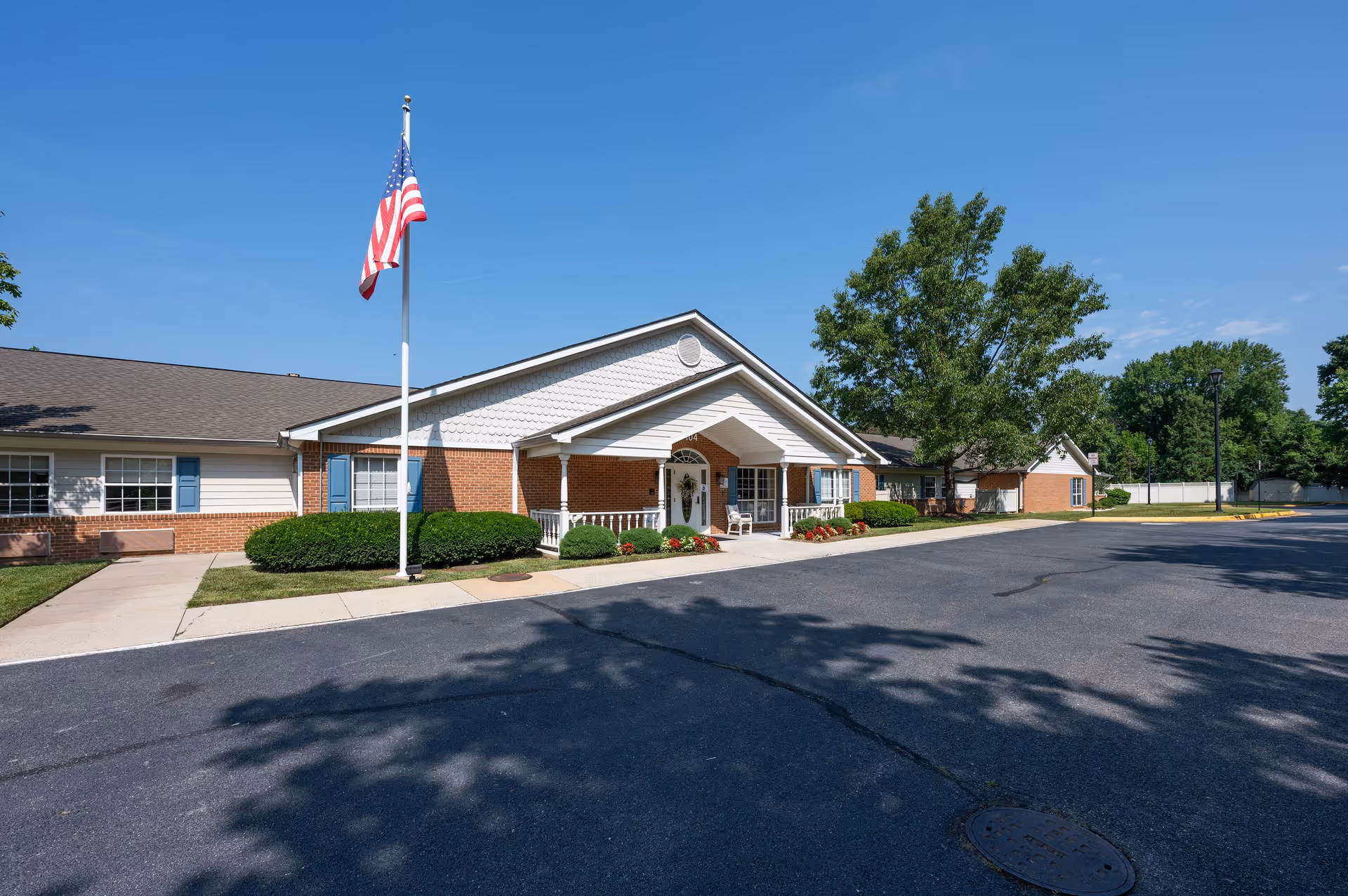 Exterior front view of a single-story brick and siding building with a covered entrance, an American flag on a flagpole, well-maintained bushes, and a paved driveway under a clear blue sky.