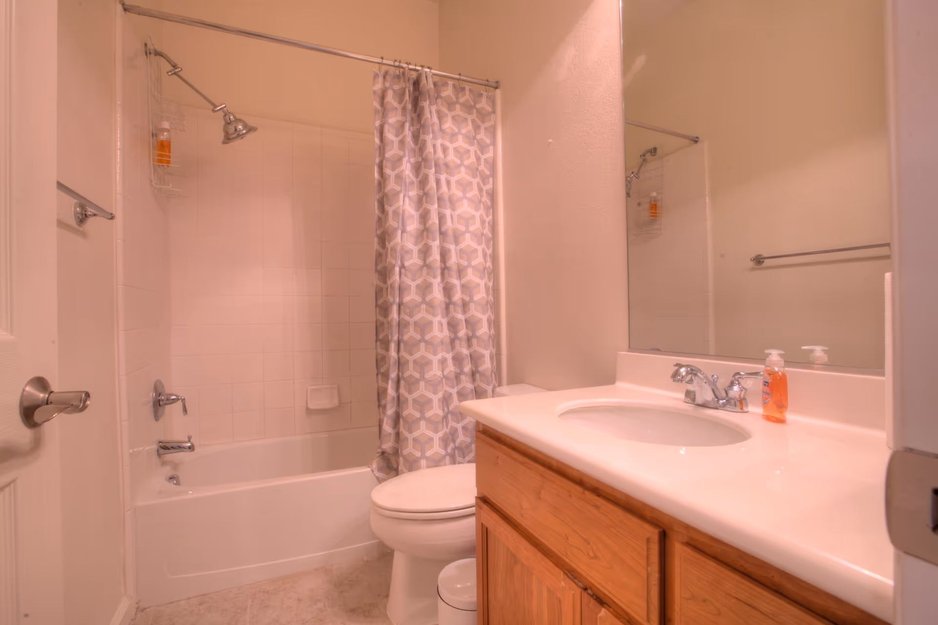 A bathroom with a white bathtub and shower combination, a patterned shower curtain, a toilet, a wooden vanity with a white countertop and sink, a large mirror above the sink, and a bottle of hand soap on the countertop.
