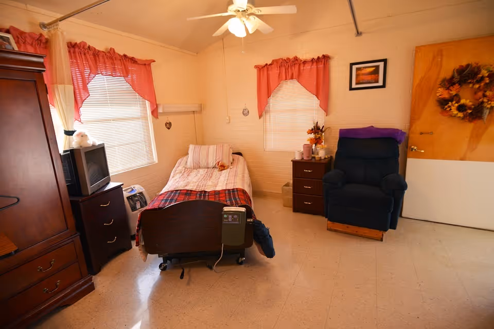 A small, cozy nursing facility bedroom with a single bed covered in a striped and plaid blanket. The room has two windows with red curtains, a wooden armoire, a TV on a small dresser, a reclining chair, and a wooden door decorated with a fall-themed wreath. The floor is tiled, and the walls are light-colored with some decorations.