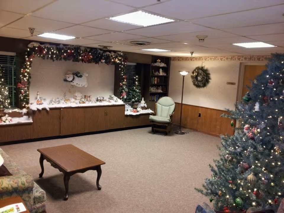 A cozy living room decorated for Christmas with a large decorated Christmas tree on the right, a wooden coffee table in the center, a green cushioned chair near a floor lamp, and a display of holiday decorations including a snowman figure and miniature houses on a cabinet against the wall. Garland with lights adorns the window area.
