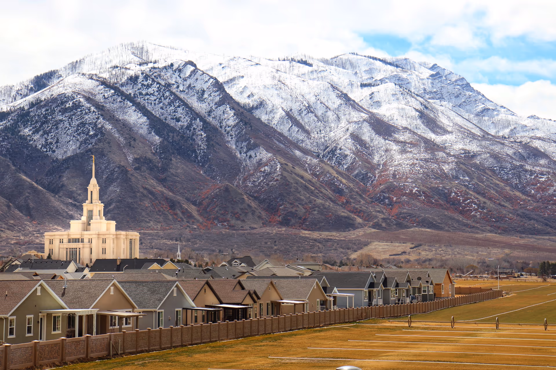 Rows of houses and a large pale temple-like building in front of snow-covered mountains under a cloudy sky.