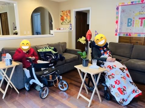 Two elderly individuals sitting in a living room area. One person is seated on a couch with a walker in front of them, and the other is in a wheelchair covered with a festive blanket. There are small tables with drinks and a plant, and a colorful birthday banner is visible on the wall behind them.