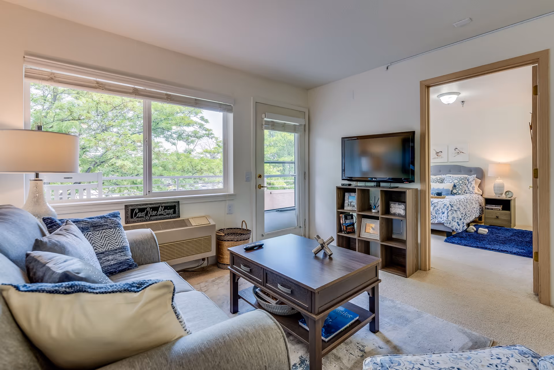 A cozy living room with a gray sofa adorned with multiple pillows, a wooden coffee table with drawers, and a TV on a cube shelf unit. The room has a large window and a glass door leading to a balcony with green trees visible outside. Through an open doorway, a bedroom with a bed, nightstand, lamp, and blue rug is visible.