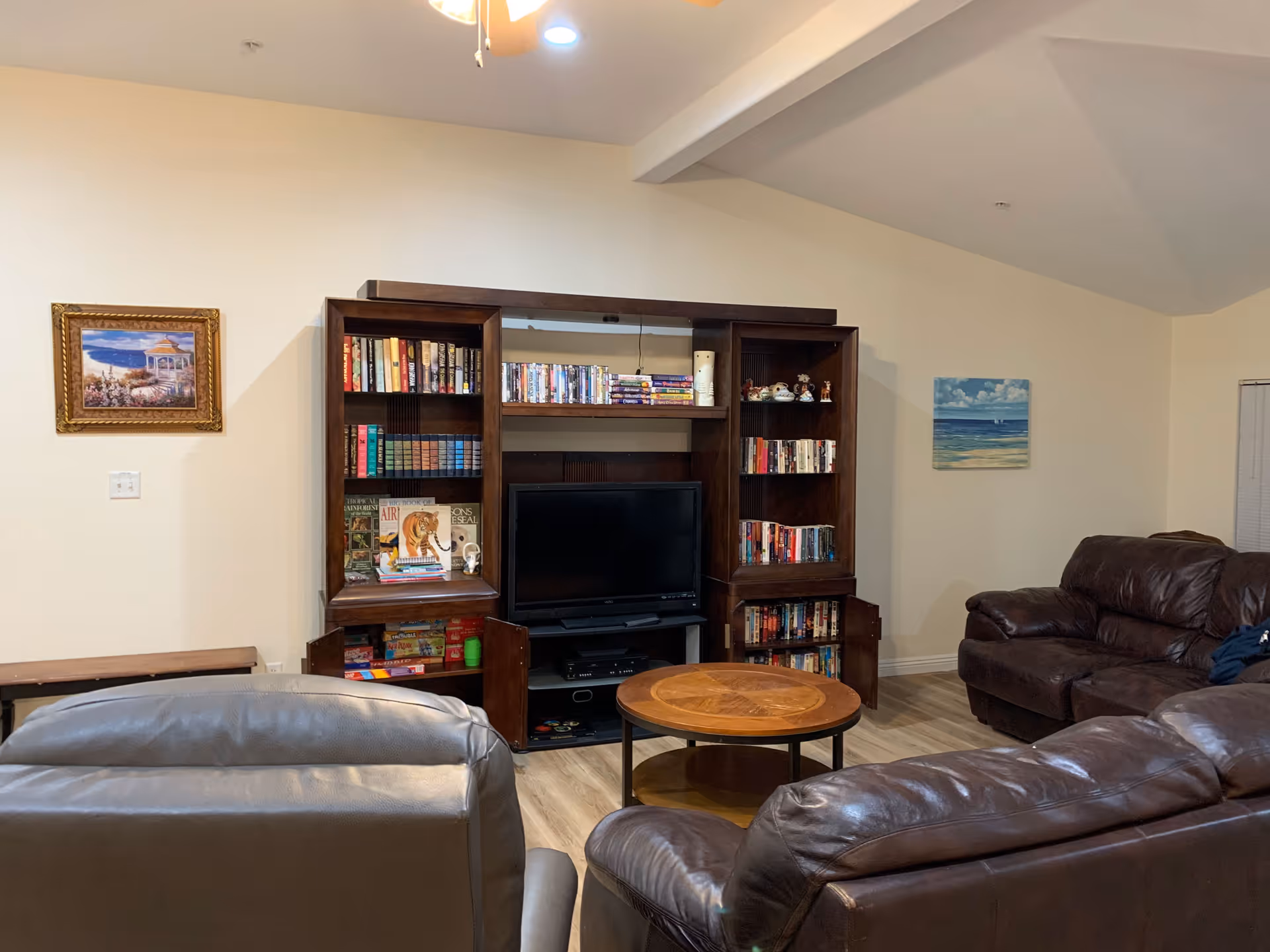 A cozy living room with brown leather sofas and a matching armchair arranged around a round wooden coffee table. Against the wall is a dark wooden entertainment center filled with books, DVDs, and decorative items, with a flat-screen TV in the center. The walls are light-colored and decorated with two paintings, one of a gazebo by the sea and another of a beach scene.