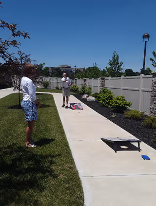 Two people playing cornhole on a sunny paved walkway next to a lawn and fence.