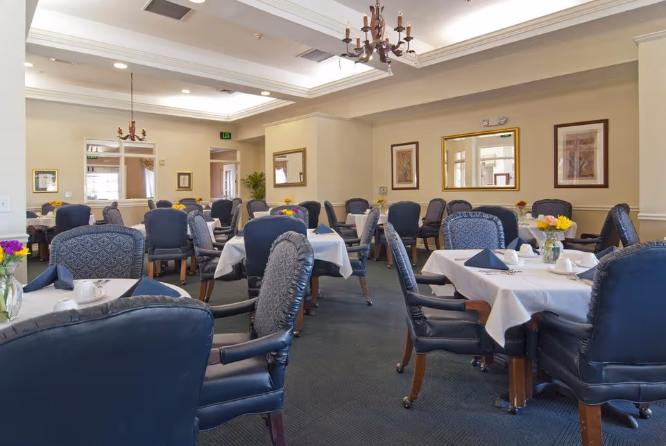 A dining room with multiple tables covered in white tablecloths, each set with blue napkins, white cups, and small flower arrangements. The room has blue upholstered chairs, beige walls with framed artwork, and a chandelier hanging from the ceiling.