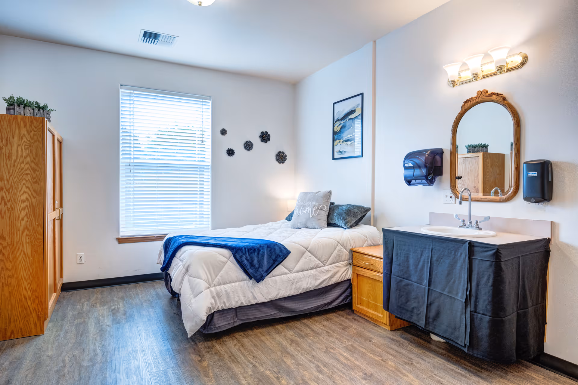 A cozy bedroom with a single bed covered in a white comforter and blue throw blanket. There is a pillow on the bed with the words 'No place like home'. The room has a wooden floor, a window with blinds, a wooden wardrobe, and a small wooden nightstand. A sink with a mirror above it and wall-mounted soap and paper towel dispensers are on the right side of the room. The walls are decorated with a framed abstract painting and metal flower wall art.