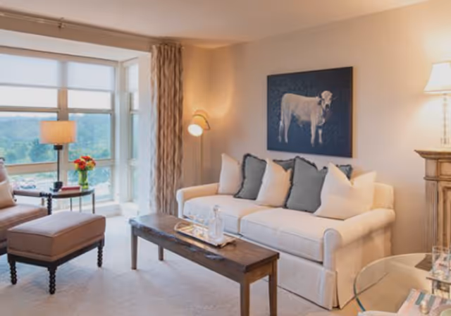 A cozy living room with a white sofa adorned with multiple pillows, a wooden coffee table with a tray and glassware, a cushioned armchair with an ottoman, a floor lamp, a table lamp on a wooden side table, and a large window with a view of greenery outside. A painting of a cow hangs on the wall above the sofa.