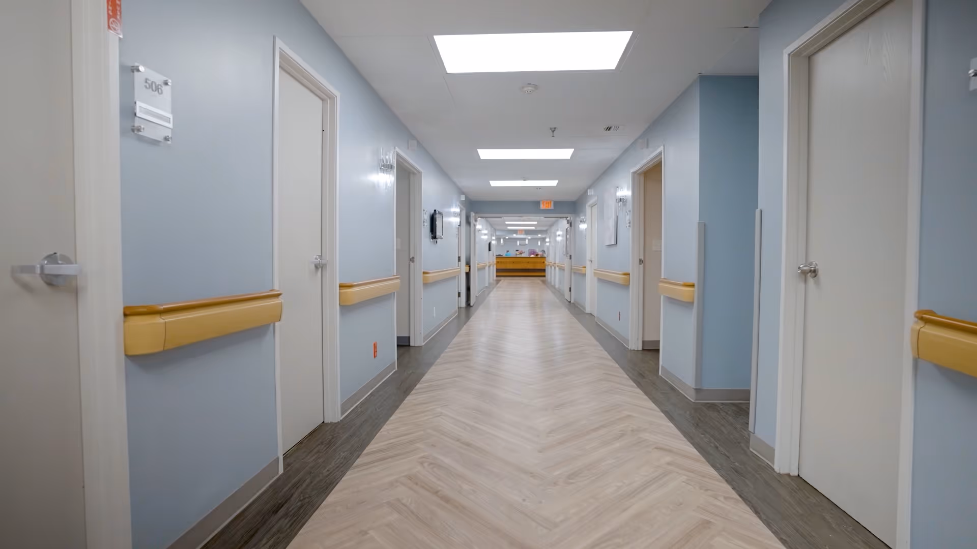 Long, well-lit interior hallway of a care facility with doors and handrails on both sides and a reception desk at the far end.