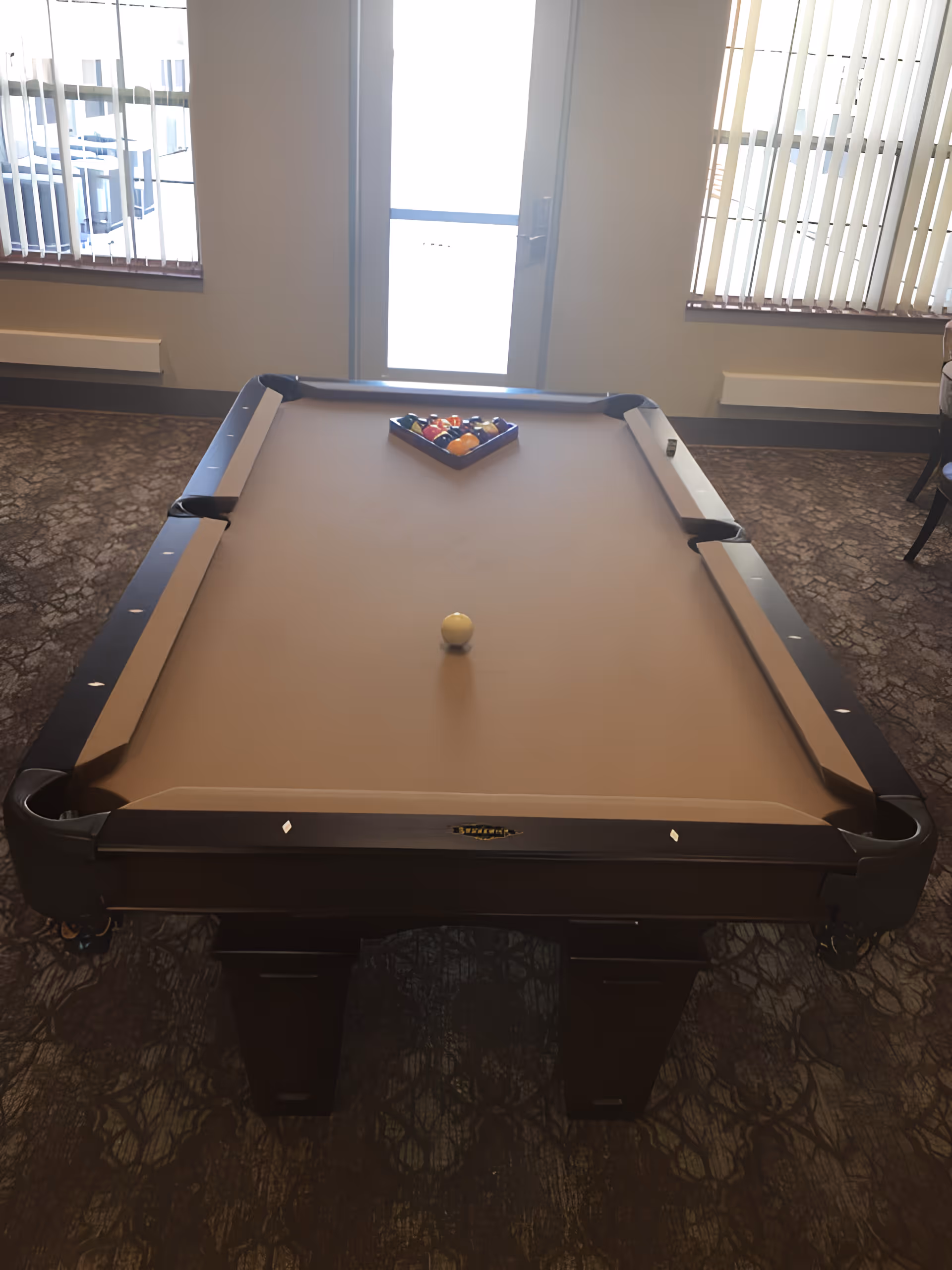 A pool table with a beige felt surface set up for a game with a triangle rack of billiard balls at one end and a cue ball near the center. The room has carpeted flooring, two windows with vertical blinds, and a door with a window letting in bright light.
