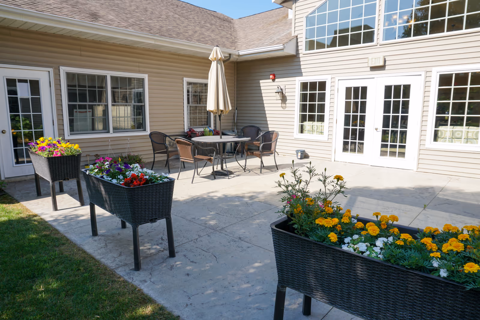 Outdoor patio area with three rectangular black planters filled with colorful flowers, a round table with four chairs, and a closed beige umbrella. The patio is adjacent to a beige building with multiple windows and glass doors.