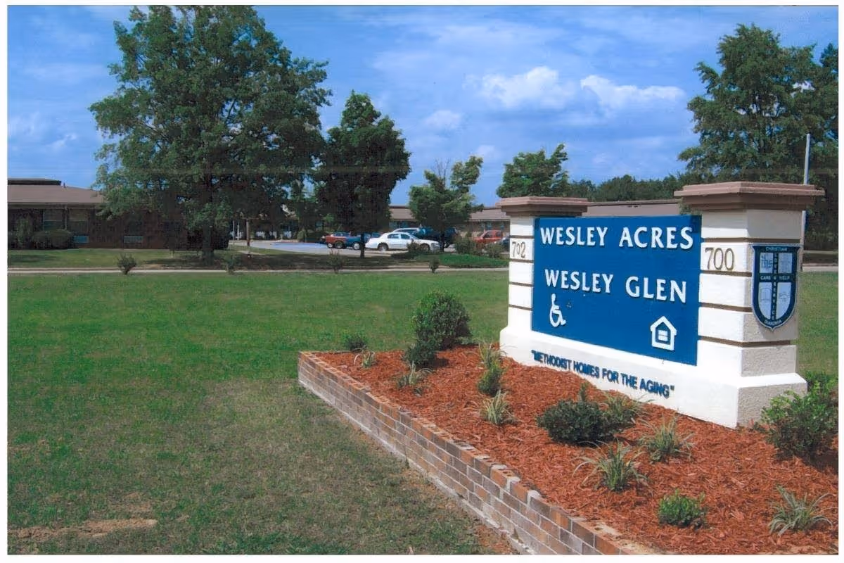 Outdoor view of a landscaped area with a large sign reading 'Wesley Acres Wesley Glen' and 'Methodist Homes for the Aging'. The sign is surrounded by mulch and small plants, with trees, grass, and parked cars in the background under a partly cloudy sky.