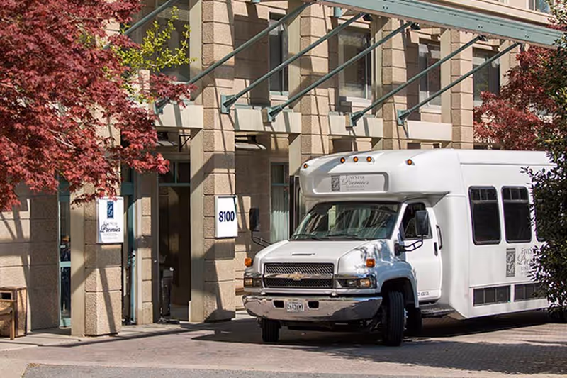 Exterior view of Five Star Premier Residences of Chevy Chase showing a beige stone building with large windows and a white shuttle bus parked in front. There are red-leaved trees on either side of the entrance and the building number 8100 is visible on a sign next to the door.